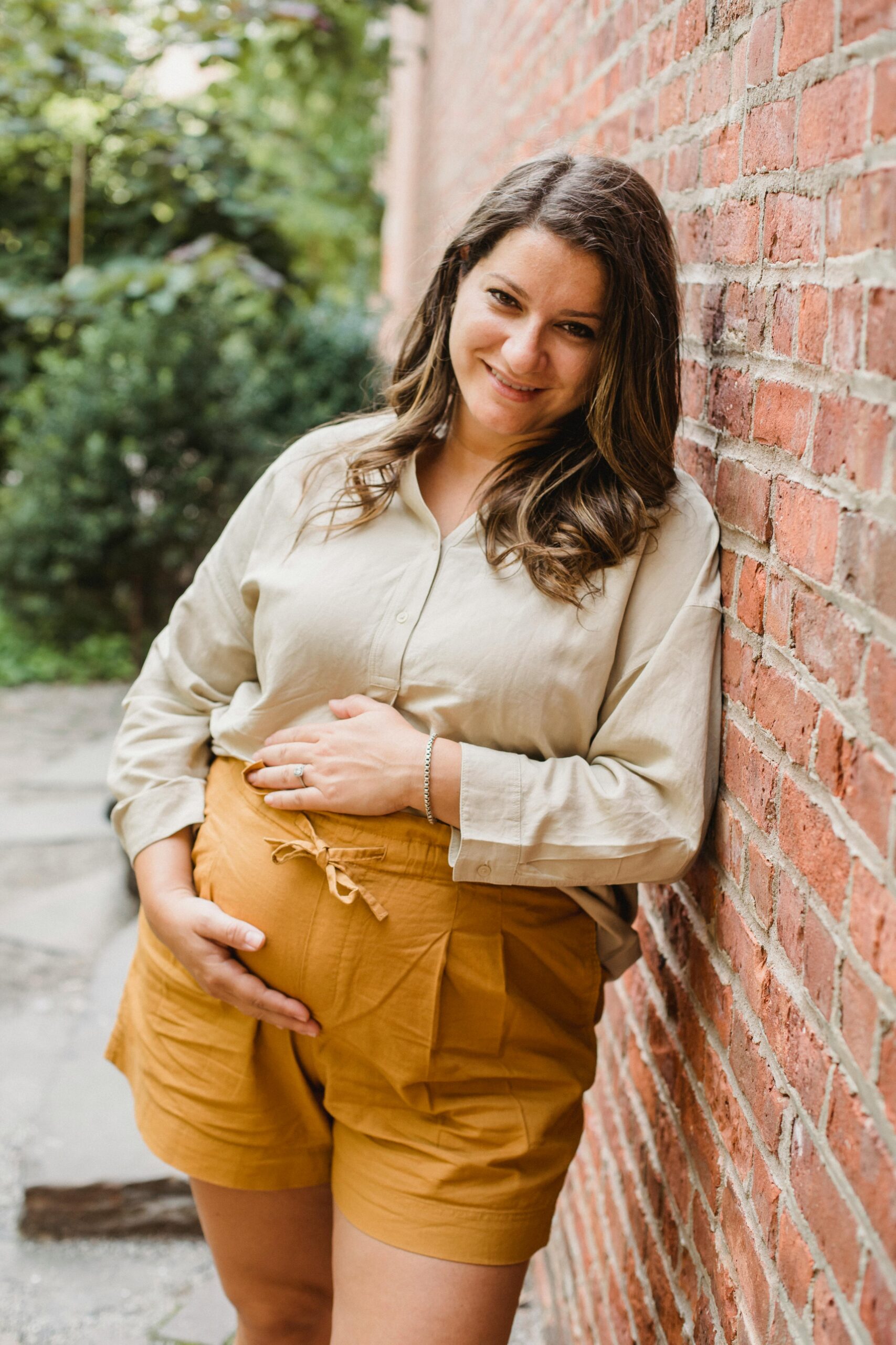 Positive pregnant female wearing shorts touching belly and leaning on brick wall while looking at camera in street on blurred background